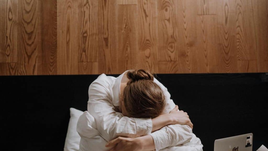 A person sitting on a bed with their head down and arms wrapped around their knees.