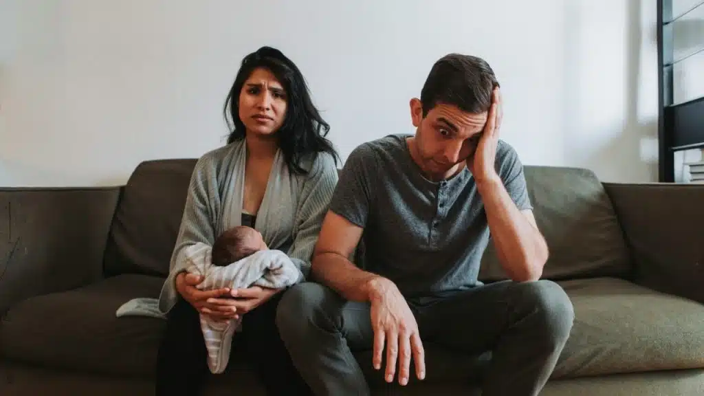 A man and woman sit on a couch; the woman holds a swaddled infant.