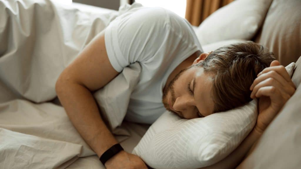 A man in a light blue T-shirt lying on a bed with white pillows