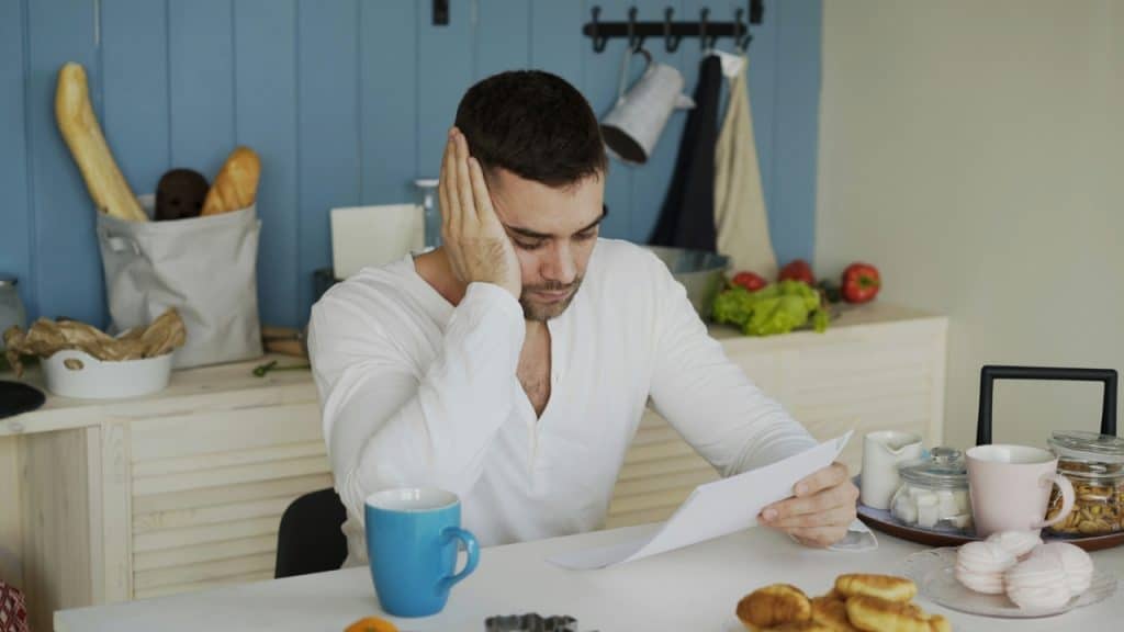 A man sits at a kitchen table holding a paper with a concerned facial expression.