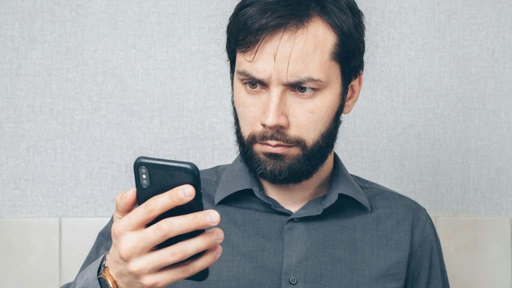 A bearded man with a serious expression looks down at his smartphone screen.