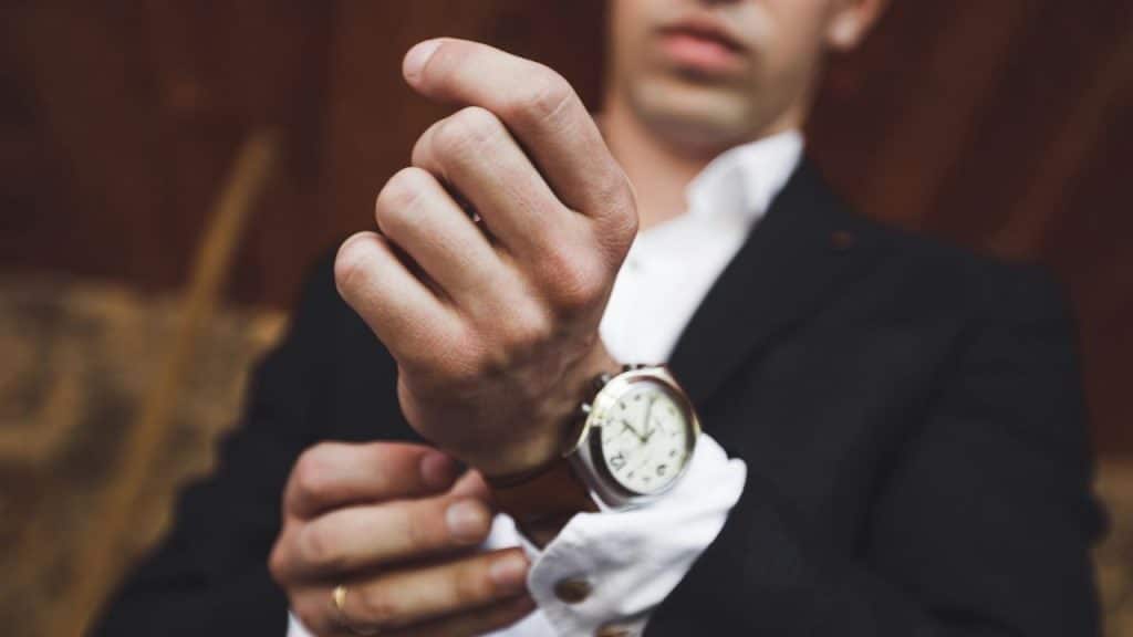 A person in a black blazer and white shirt adjusts their leather-strap wristwatch.