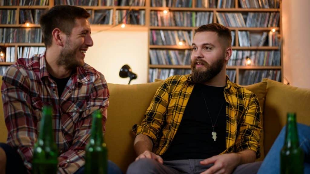 Two men in plaid shirts sit on a couch talking and drinking beer.