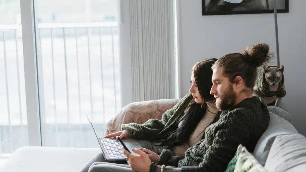 A couple sitting on a couch using a laptop and tablet while a cat sits behind them.