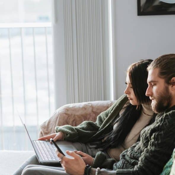 A couple sitting on a couch using a laptop and tablet while a cat sits behind them.