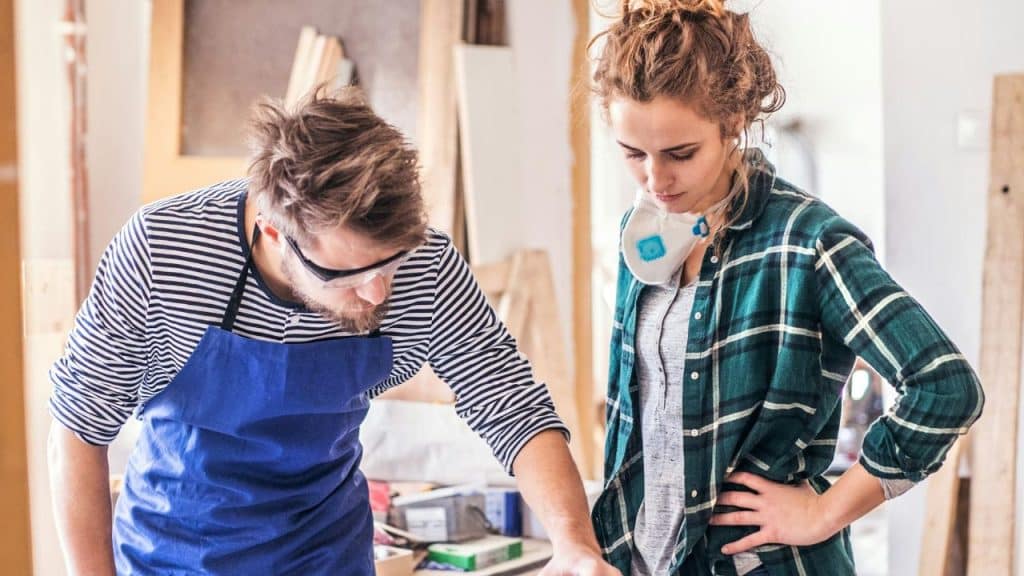 A man in an apron and safety glasses works while a woman observes him.