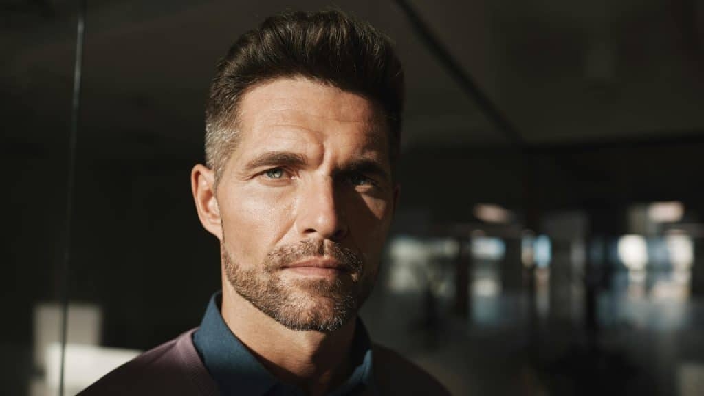A close-up portrait of a man with dark hair looking directly at the camera.