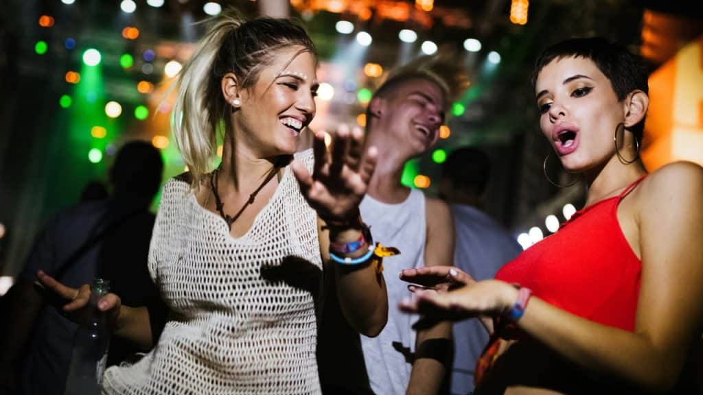 Two women dance and laugh at a crowded nightclub with bright green and orange lights.