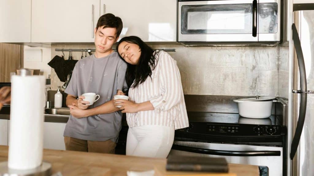 A couple standing in a kitchen, holding mugs and leaning together.