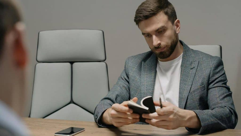 A man in a blazer reviewing notes at a desk during a meeting.