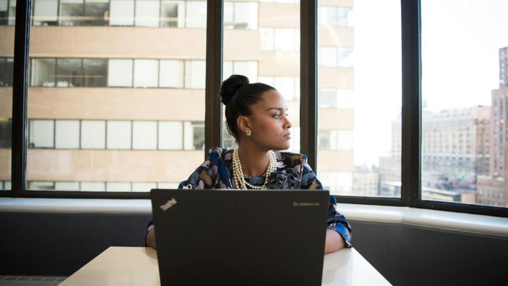A woman sitting at a table with a laptop, looking out a window.