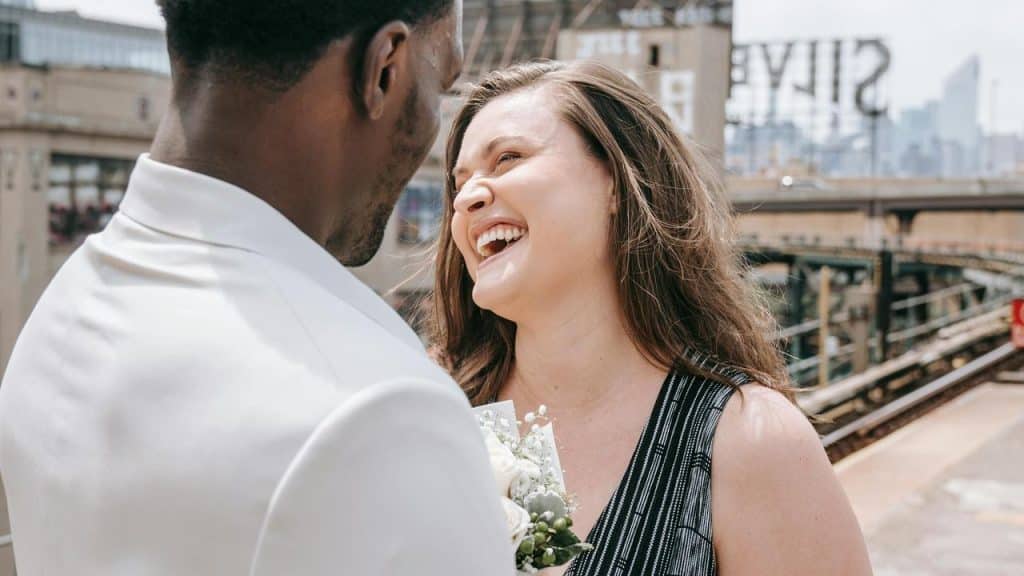A couple smiling and talking outdoors near train tracks.