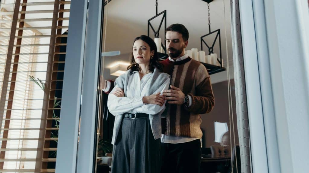 A man in a striped sweater stands behind a woman with her arms crossed indoors.