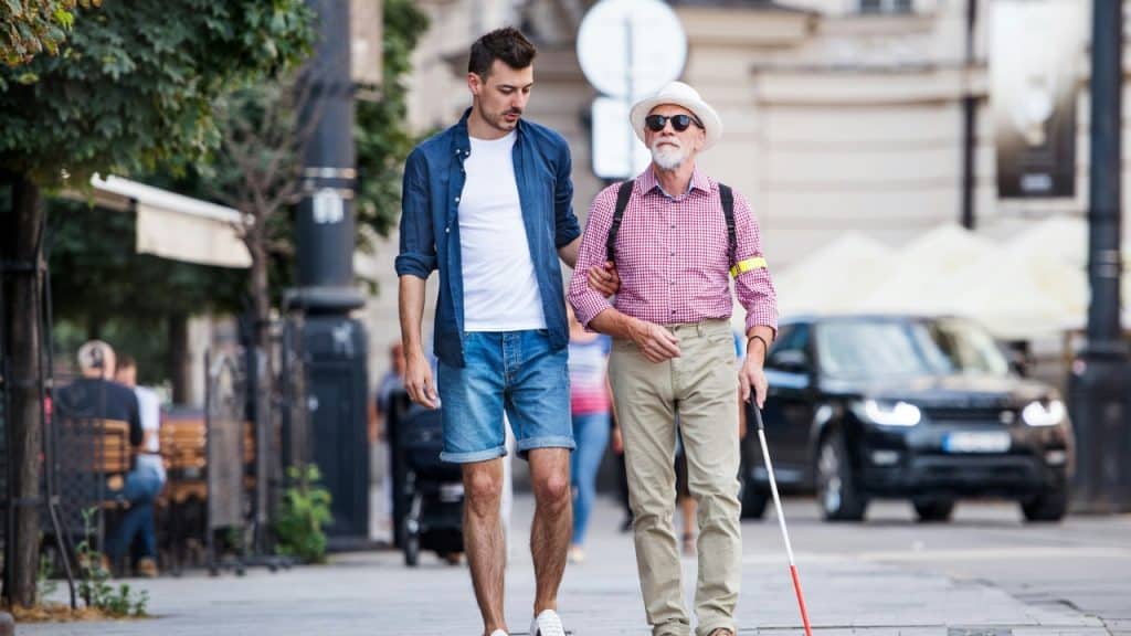 A young man walks alongside an older man holding a white cane outdoors.
