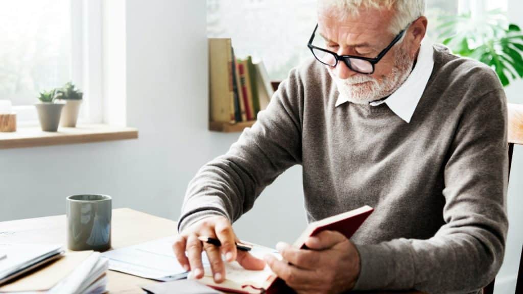 An older man with glasses and a beard writes in a notebook at a desk.