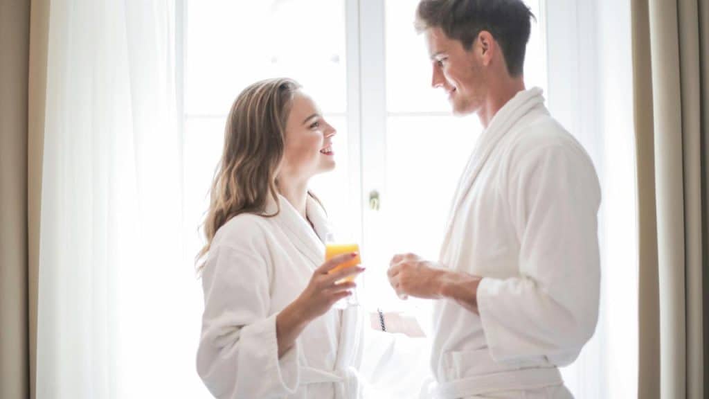 A smiling couple in white bathrobes sharing a drink in a bright room.