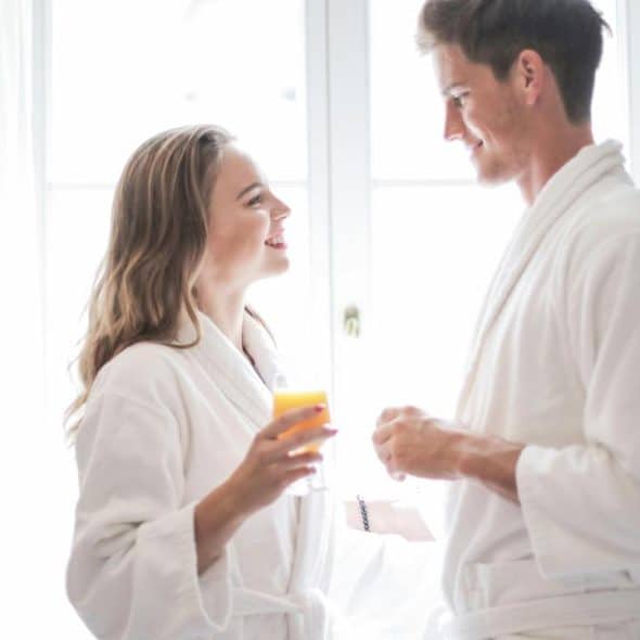 A smiling couple in white bathrobes sharing a drink in a bright room.