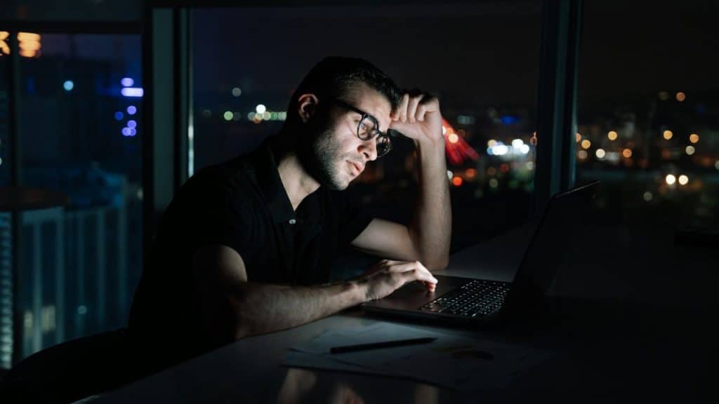 A man wearing glasses works on a laptop in a dark room at night.