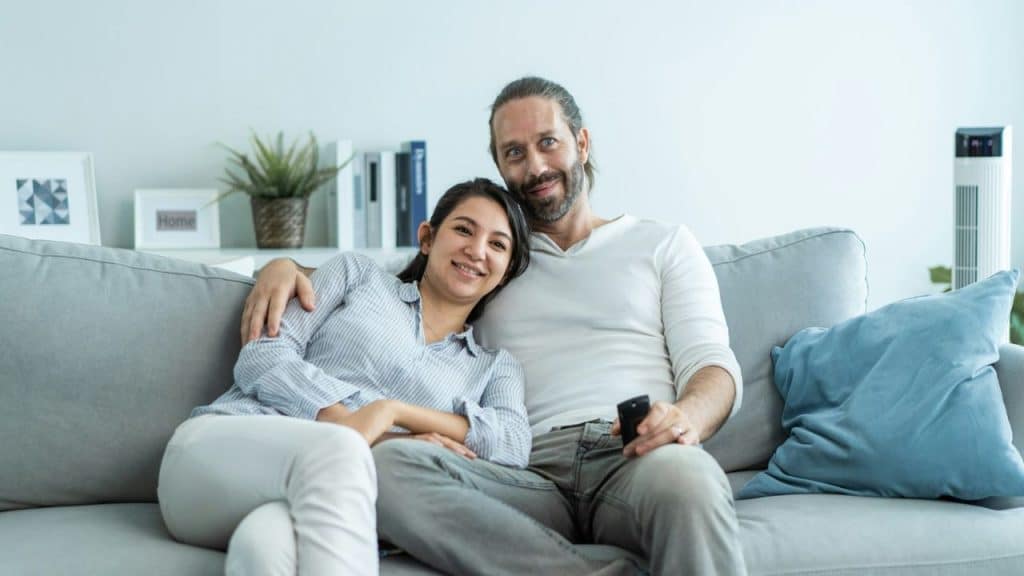 A man and woman sit together on a gray sofa, the man holding a remote.