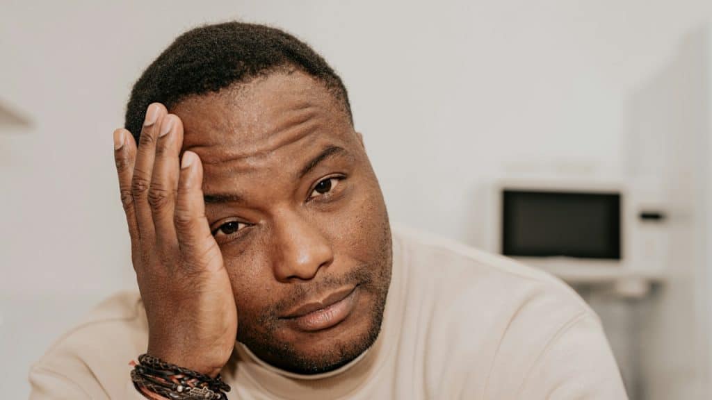 A man resting his head on his hand with a tired expression, wearing a beige shirt and bracelets.