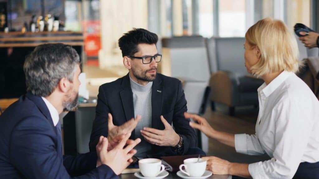 Three people sit around a small table with coffee cups, gesturing during a serious discussion.