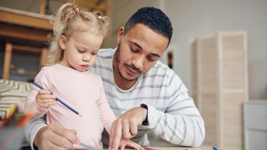A father helping his young daughter draw at a table.
