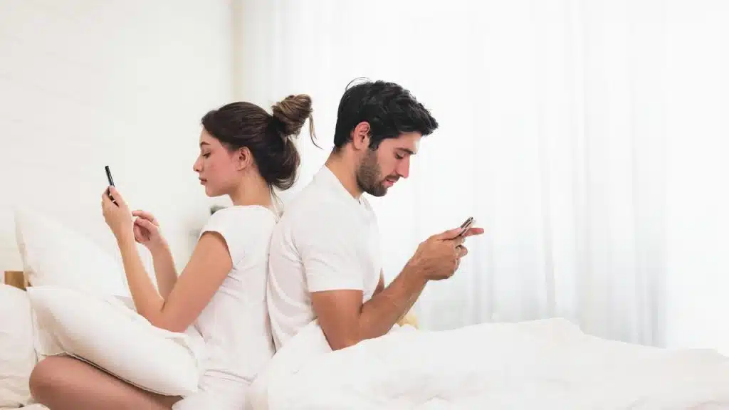 A couple sitting back to back in bed using their phones.