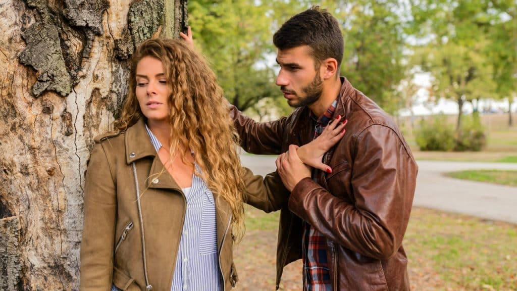 A woman leans against a tree while a man places a hand on her shoulder.