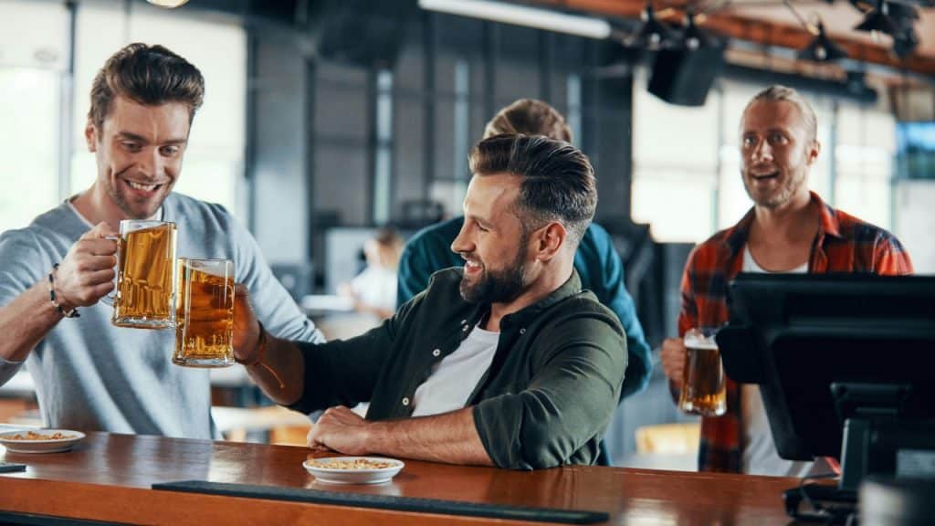 Three men stand at a wooden bar counter holding large glass mugs of beer.