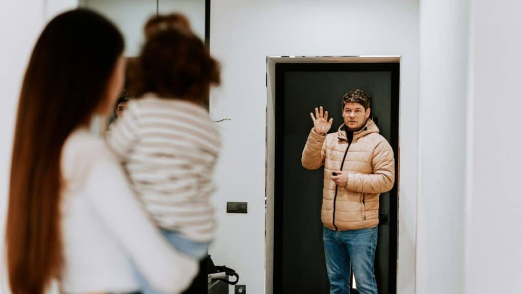 A man in a tan jacket waves goodbye while a woman holds a small child.