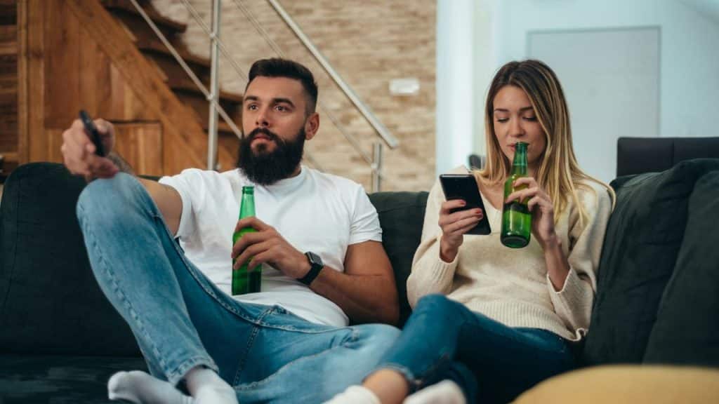 A man holds a remote and bottle while a woman uses a phone and drinks.