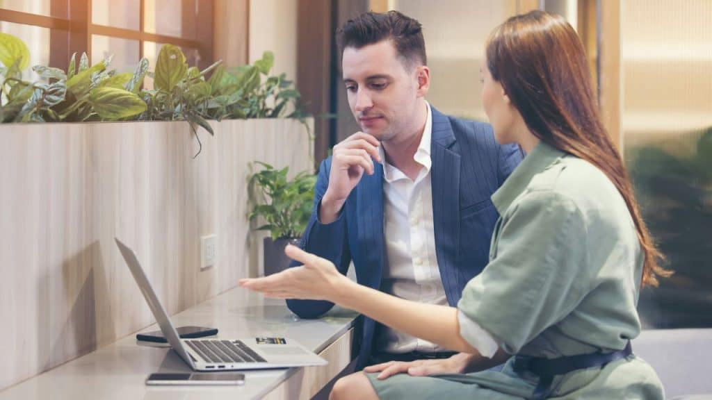 A woman gestures toward a laptop screen while a man in a suit looks on thoughtfully.