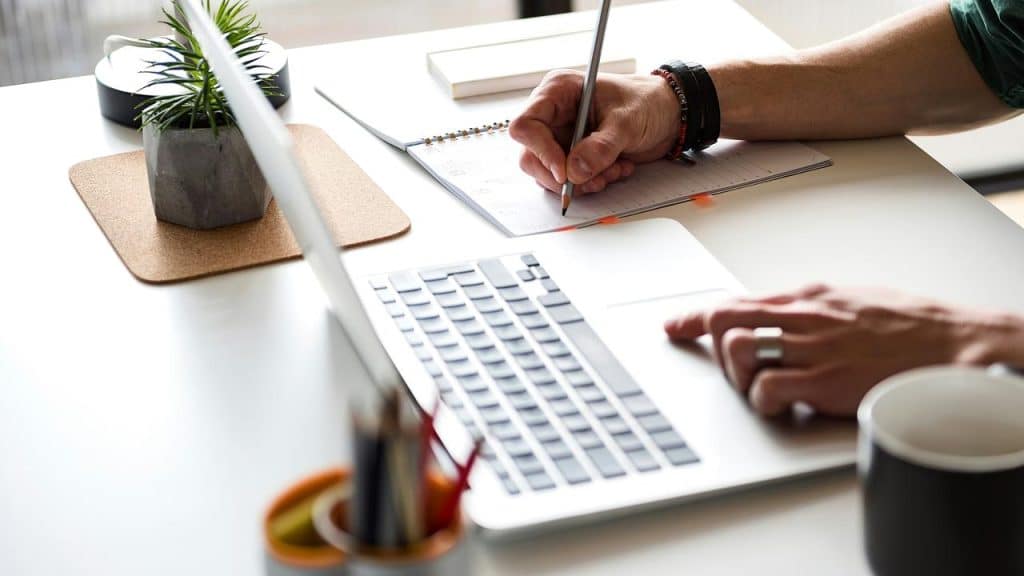 A person writing in a notebook while working on a laptop.