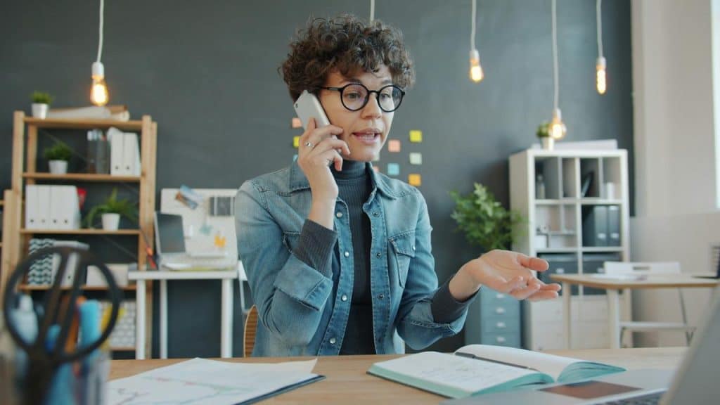 A woman talking on the phone while sitting at a desk with papers and a notebook.