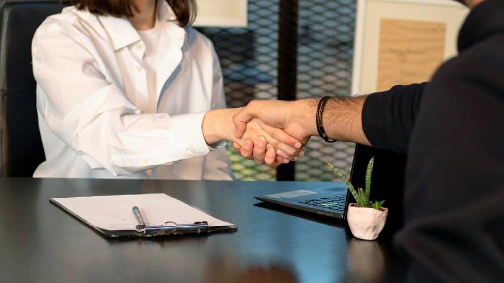 A close-up of two people shaking hands across a desk with a laptop and documents.