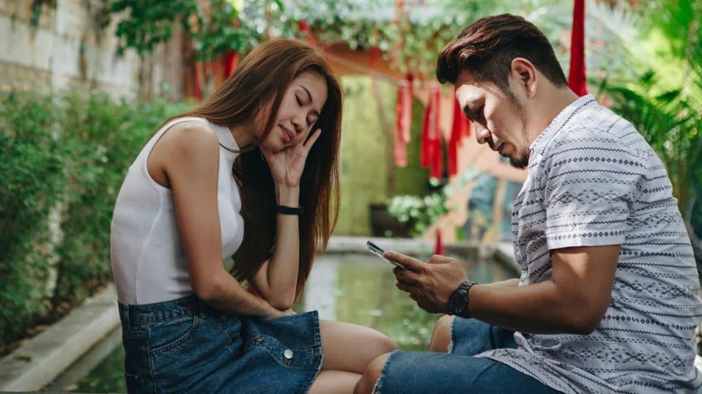 A man looks at a smartphone while a woman rests her head on her hand nearby.