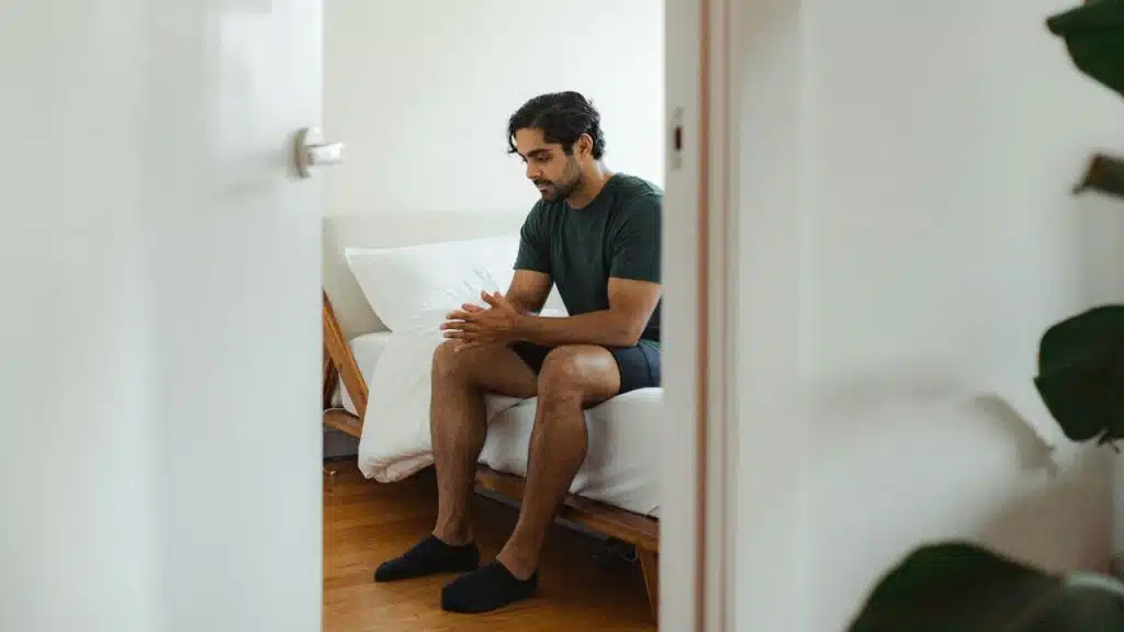 A man sits on the edge of a bed with his hands together, looking down.