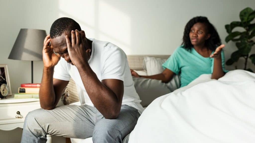 A man sits with head in hands while a woman speaks behind him in bed.