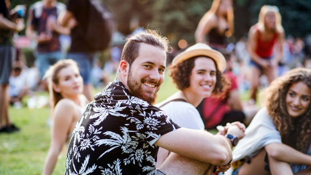 A group of cheerful young friends is sitting on the ground at a summer festival, looking at the camera.
