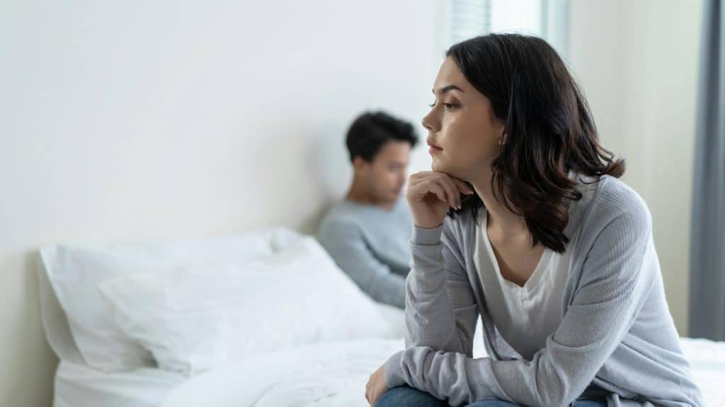 A young woman sits on the edge of a bed while a man sits away from her in the background.