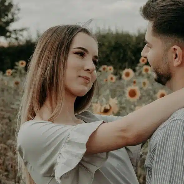 A woman staring in a man’s eyes while resting her arms on his arms as they stand in a sunflower field.