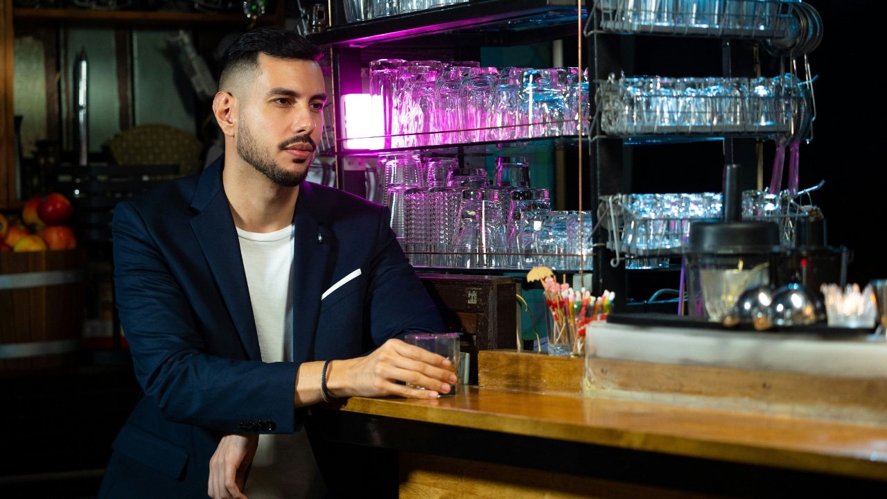 A man in a blue blazer sits alone at a bar holding a drink near glass shelves.
