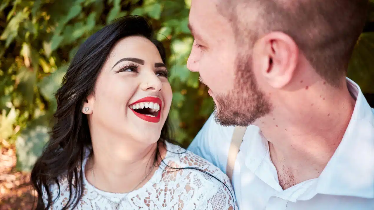 A woman with red lipstick laughs while looking up at a man with a beard.