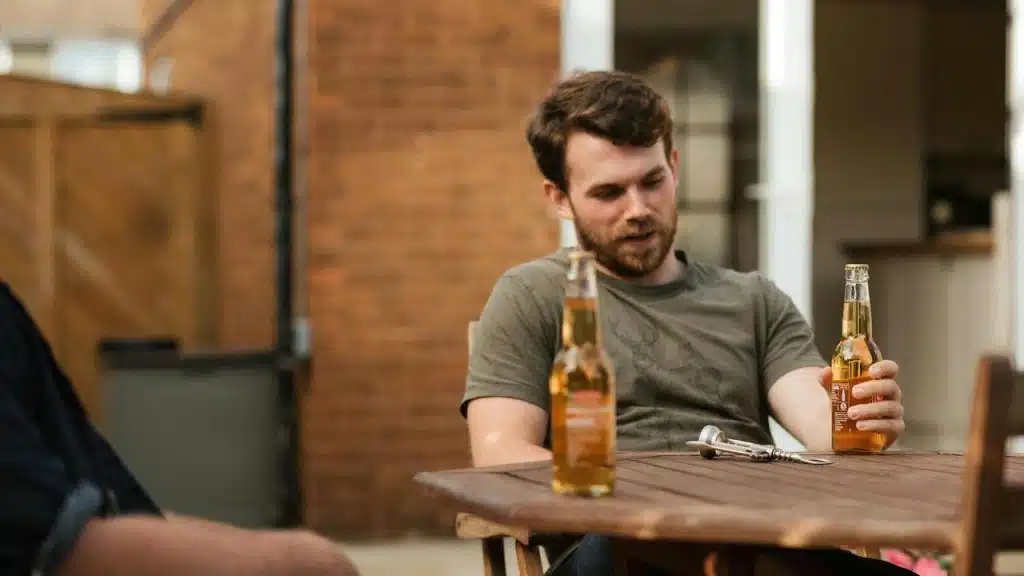 A man sits outdoors holding a beer bottle at a wooden table.