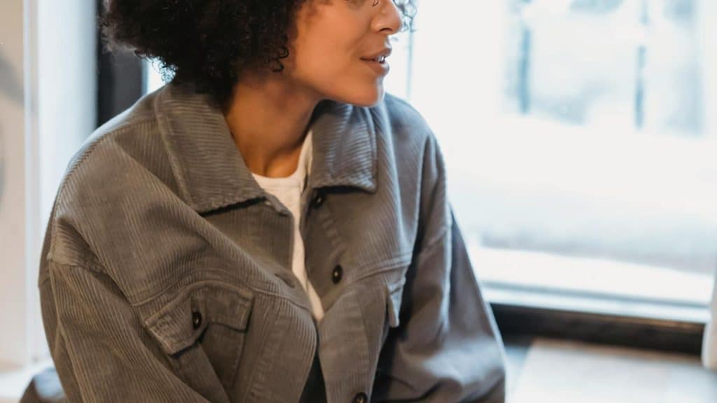 A woman in a gray jacket sitting by a window and talking.