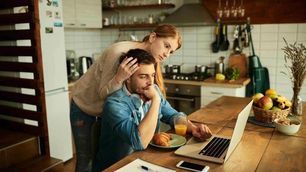 A woman leans over a man’s shoulder while he sits at a table using a laptop.