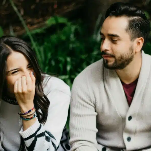A woman laughing with her hand over her face sitting next to a bearded man.