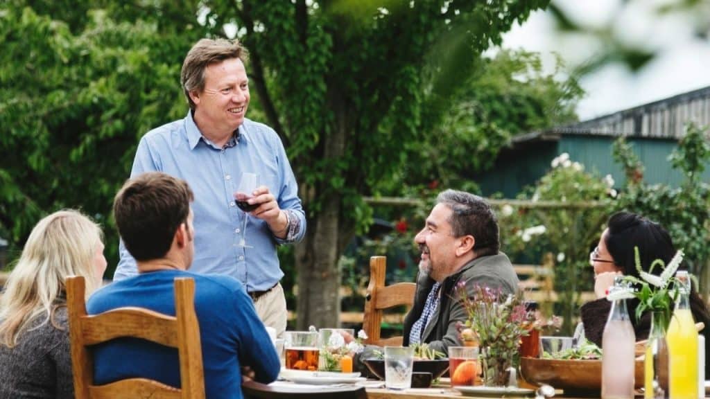 A man stands holding a glass of wine, talking to a group seated at an outdoor table.