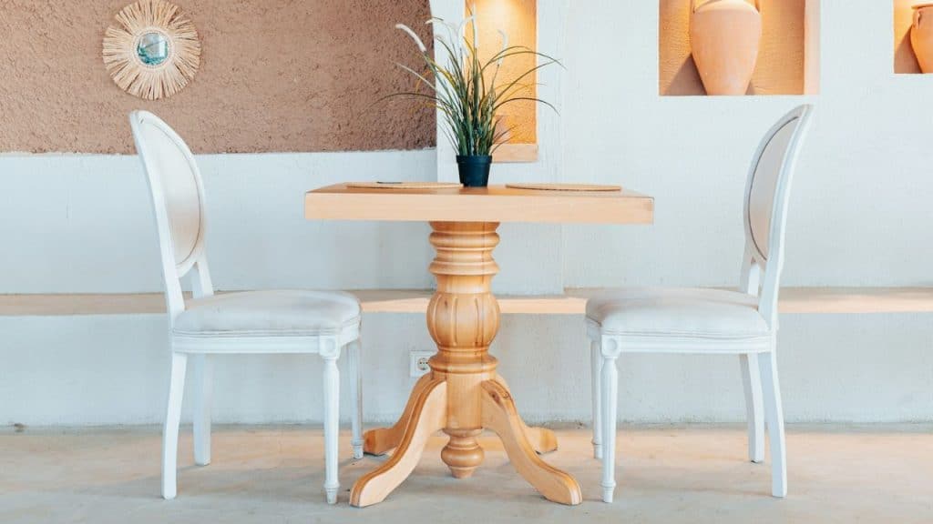 Two empty white chairs face an ornate wooden table with a small potted plant on top.