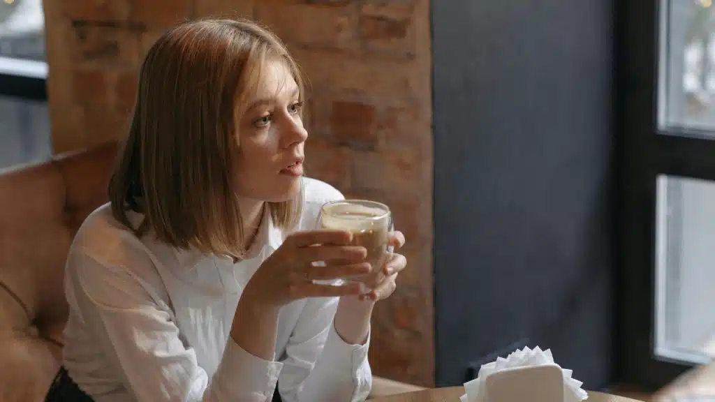 A woman sits in a cafe holding a glass of coffee while looking out of a window.